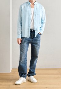 Camicia a maniche lunghe in lino azzurro chiaro sopra una canotta bianca, abbinata a jeans dritti blu e sneakers bianche. Design semplice, vestibilità casual.