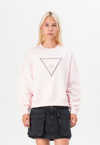 Pink sweatshirt with crew neckline, loose fit, and a triangular rhinestone accent on the front. Worn with a black denim skirt.