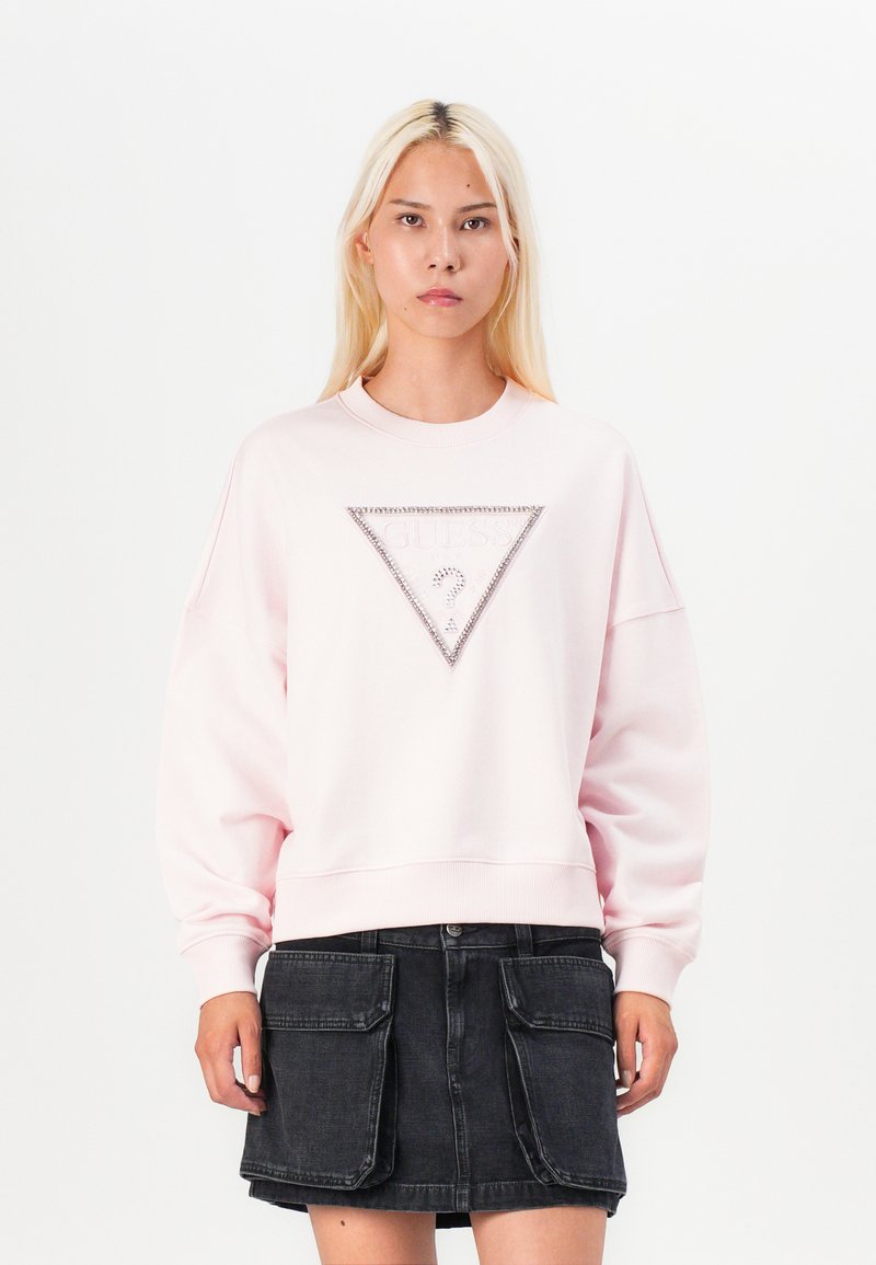 Pink sweatshirt with crew neckline, loose fit, and a triangular rhinestone accent on the front. Worn with a black denim skirt.