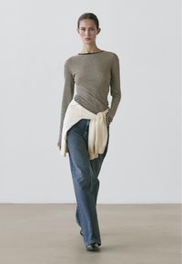 Langärmliges gestreiftes Oberteil mit Rundhalsausschnitt, kombiniert mit weit geschnittenen blauen Jeans und einem hellen Pullover, der um die Taille gelegt ist. Schwarze Schuhe.