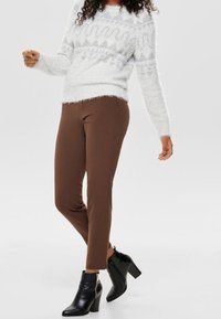 Bruna, figurnära byxor i kombination med en vit, texturerad tröja med geometriskt mönster; svarta stilettklackar fulländar outfiten.