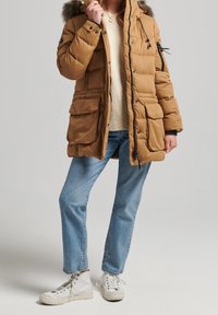 Veste matelassée marron avec une capuche bordée de fourrure, fermeture éclair sur le devant, plusieurs poches avant, et poignets élastiques, associée à un jean bleu clair et des baskets blanches.