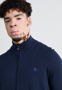 Suéter azul marino con cremallera y un alto cuello, textura de punto y pequeño logotipo de árbol azul en el lado izquierdo del pecho. Diseño ajustado.