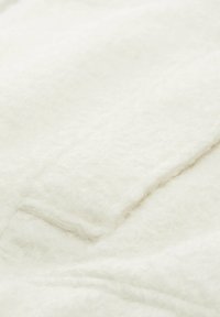 Weißer, flauschiger Stoff mit einer weichen Textur, der einen subtilen Glanz aufweist. Das Bild konzentriert sich auf einen Abschnitt mit einem Taschendetail und hebt seine Wärme hervor.