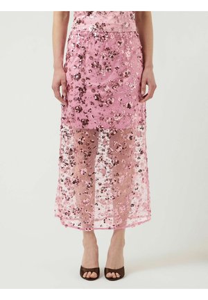 Kvinde iført en pink blomsterbesat midi-nederdel med gennemsigtigt lag og brune åbne hæle, stående mod en ensfarvet baggrund.