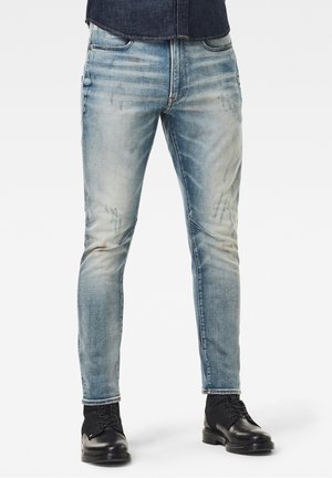 Homme portant un jean slim bleu délavé et des bottes en cuir noir à lacets, debout devant un fond uni.
