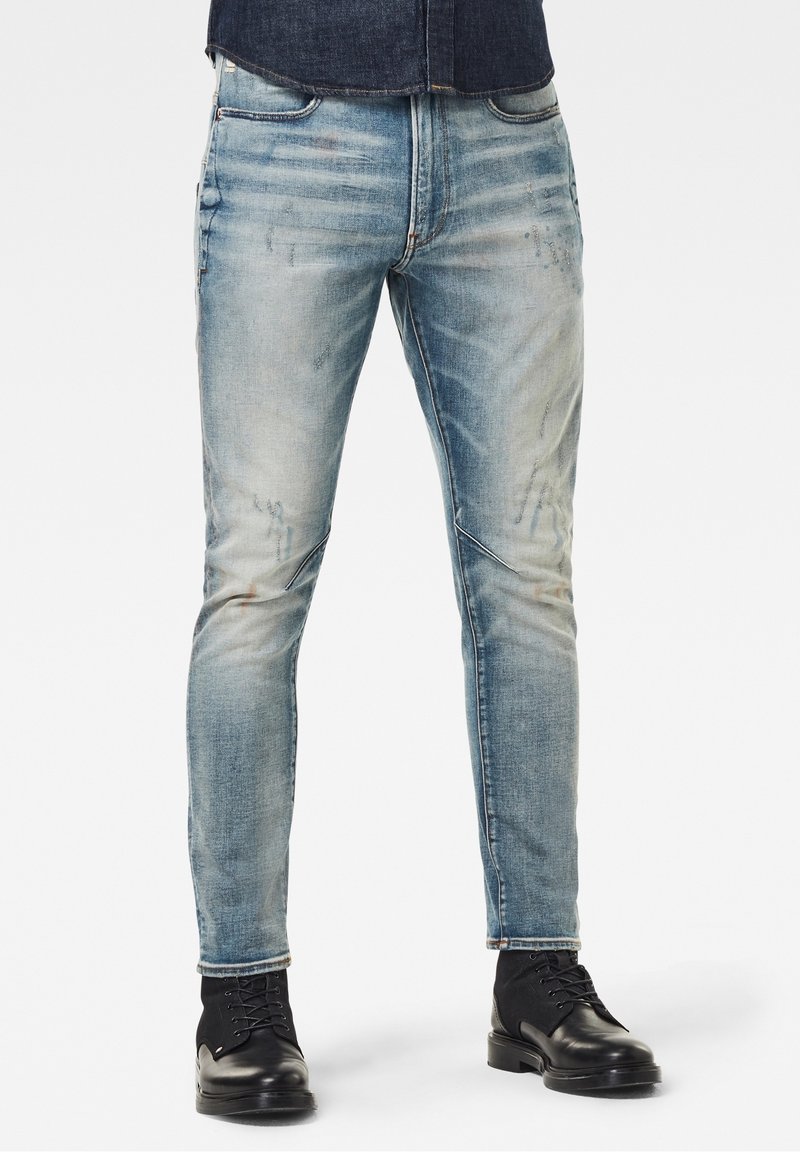 Homme portant un jean slim bleu délavé et des bottes en cuir noir à lacets, debout devant un fond uni.