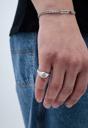 Strieborný prsteň s okrúhlym tvarom a podrobným kruhovým gravírovaním, nosený na ruke spolu so strieborným reťazovým náramkom, s viditeľnou modrou džínsovinou.