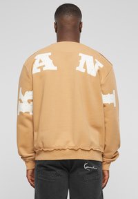 Beige sweatshirt med store, slidte hvide bogstaver på ryggen og ærmerne. Ribbet manchetter og kant med rå kantdetaljer.