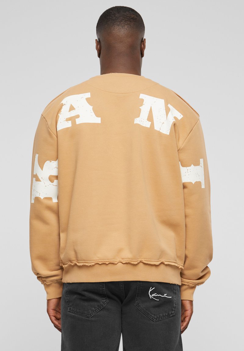 Beige sweatshirt med store, slidte hvide bogstaver på ryggen og ærmerne. Ribbet manchetter og kant med rå kantdetaljer.