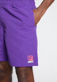 Violetit shortsit, joissa on joustava vyötärönauha, sivutaskut ja punainen-oranssi kuva-aihe vasemmassa lahkeessa. Sileä, kevyt kangas.