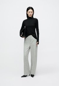 Svart långärmad polotröja, ljusgrå vida byxor och spetsiga svarta klackar. Modellen håller en svart kuvertväska. Enkel, modern design.