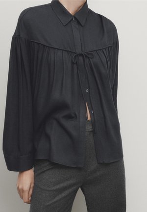 Zwarte blouse met lange mouwen met geplooide inzet en strikdetail, gedragen met hooggetailleerde donkergrijze broek.