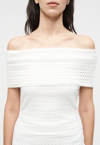 Γυναίκα με λευκό πλεκτό τοπ χωρίς ώμους, με ακάλυπτους ώμους και άνω μέρος στήθους σε μονόχρωμο φόντο.