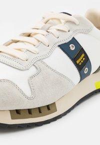 Sneaker da uomo con rete bianca, dettagli in suede beige, marchio blu e giallo, e suola testurizzata. Presenta un design con lacci e modelli unici.