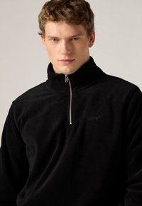 Μαύρο πουλόβερ από φλις με ψηλό γιακά, ημισεληνοειδές φερμουάρ, απαλό υλικό και διακριτικό ανάγλυφο λογότυπο στο στήθος.