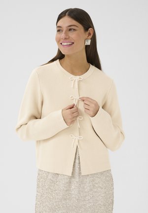 Beige gestrickte Strickjacke mit rundem Ausschnitt, drei mit Perlen verzierte Schleifenverschlüsse und lockerer Passform, getragen zu einem schimmernden, hellbeigen Rock.