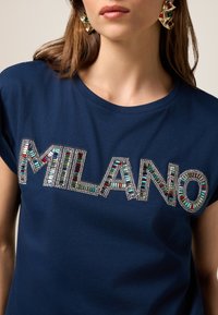 Tricou bleu marin, prezentând un logo "MILANO" împodobit cu cristale, cu pietre colorate vibrante în diferite dimensiuni și modele geometrice.