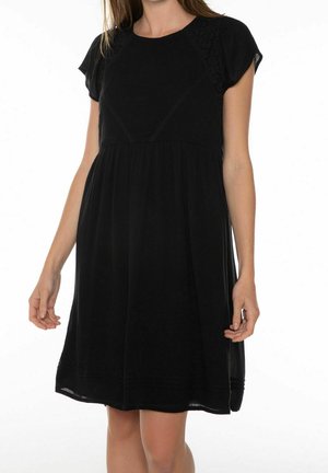 Femme portant une robe noire à manches courtes, longueur au genou, avec un col rond et des détails délicats en dentelle sur les épaules.