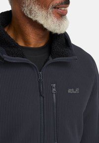 Giacca in pile blu scuro con texture a coste, chiusura con zip e colletto alto. Presenta un logo sul petto e una fodera morbida per il comfort.