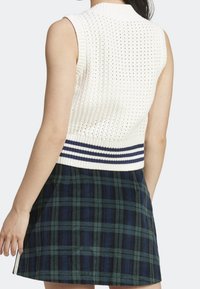 Pletená svetrová vesta v bílé barvě s texturovaným vzorem, s žebrovaným výstřihem a navy proužkovaným spodním lemem. Nošena přes kostkovanou sukni.