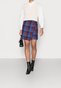 Beige gebreide mouwloze trui over een witte blouse met kraag, gecombineerd met een blauw en bordeaux geruit minirok en zwarte enkellaarsjes.