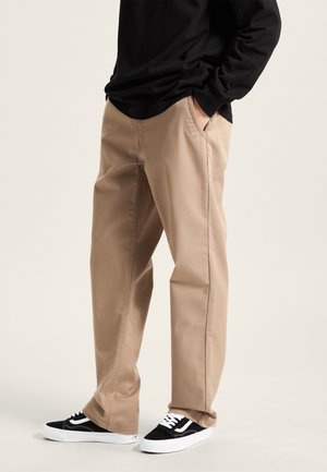 Person iført beige bukser med lige ben, sort langærmet skjorte og sorte-hvide sneakers, stående med hænderne i lommerne.