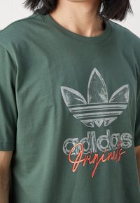 Tmavě zelené bavlněné tričko se zašlými stříbrnými logem Adidas a červeným textem "Originals", s kulatým výstřihem a krátkými rukávy.
