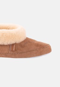 Bruna mockasiner i mockaläder med en mjuk krämfärgad fårskinnskant. Har en mjuk sula och sidov brandingdetalj. Slät yta över hela ytan.