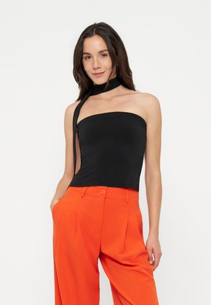 Jeune femme aux longs cheveux foncés porte un top noir sans bretelles avec un nœud au cou et un pantalon large taille haute orange vif, les mains dans les poches.