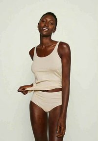 Woman wearing beige tank top and matching high-waist underwear, smiling and pulling the hem of the top slightly upward.