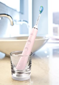 Brosse à dents électrique avec un manche rose, une tête blanche et des poils bleu-vert, placée dans un verre transparent sur une surface en marbre.