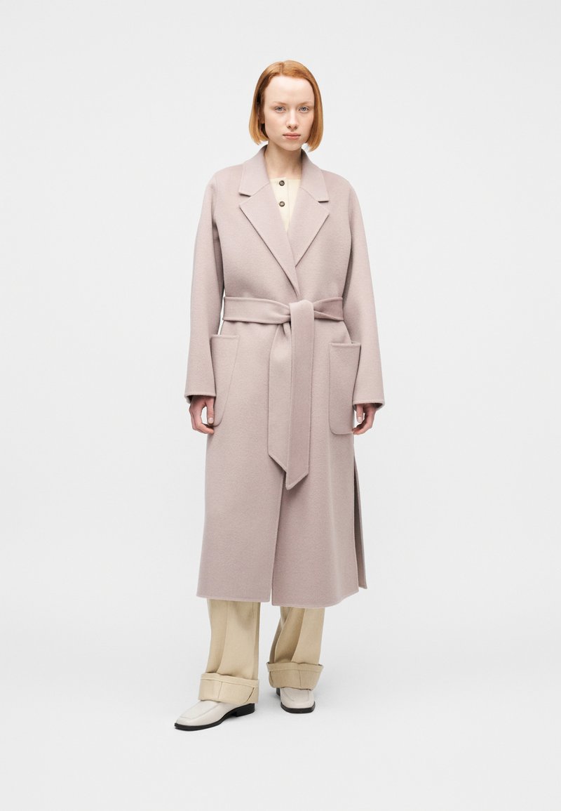 Γυναίκα με κοντά κόκκινα μαλλιά που φοράει ένα μ ακρύ μπεζ παλτό με ζώνη, εκρού πουκάμισο, φαρδιά μπεζ παντελόνια και λευκά παπούτσια, στέκεται σε λευκό φόντο.