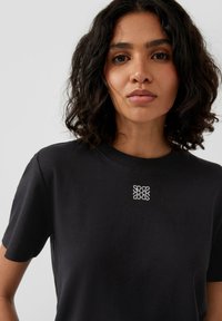 Schwarzes Baumwoll-T-Shirt mit einem strukturierten quadratischen Design und kleinen weißen Akzenten auf der Brust, rundem Ausschnitt und kurzen Ärmeln.