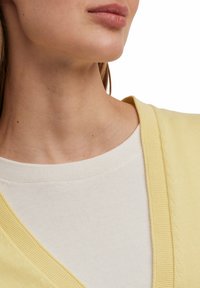 Een gele cardigan met geribbelde randen, gedragen over een witte top. De cardigan heeft een V-hals ontwerp en een gestructureerde stof.
