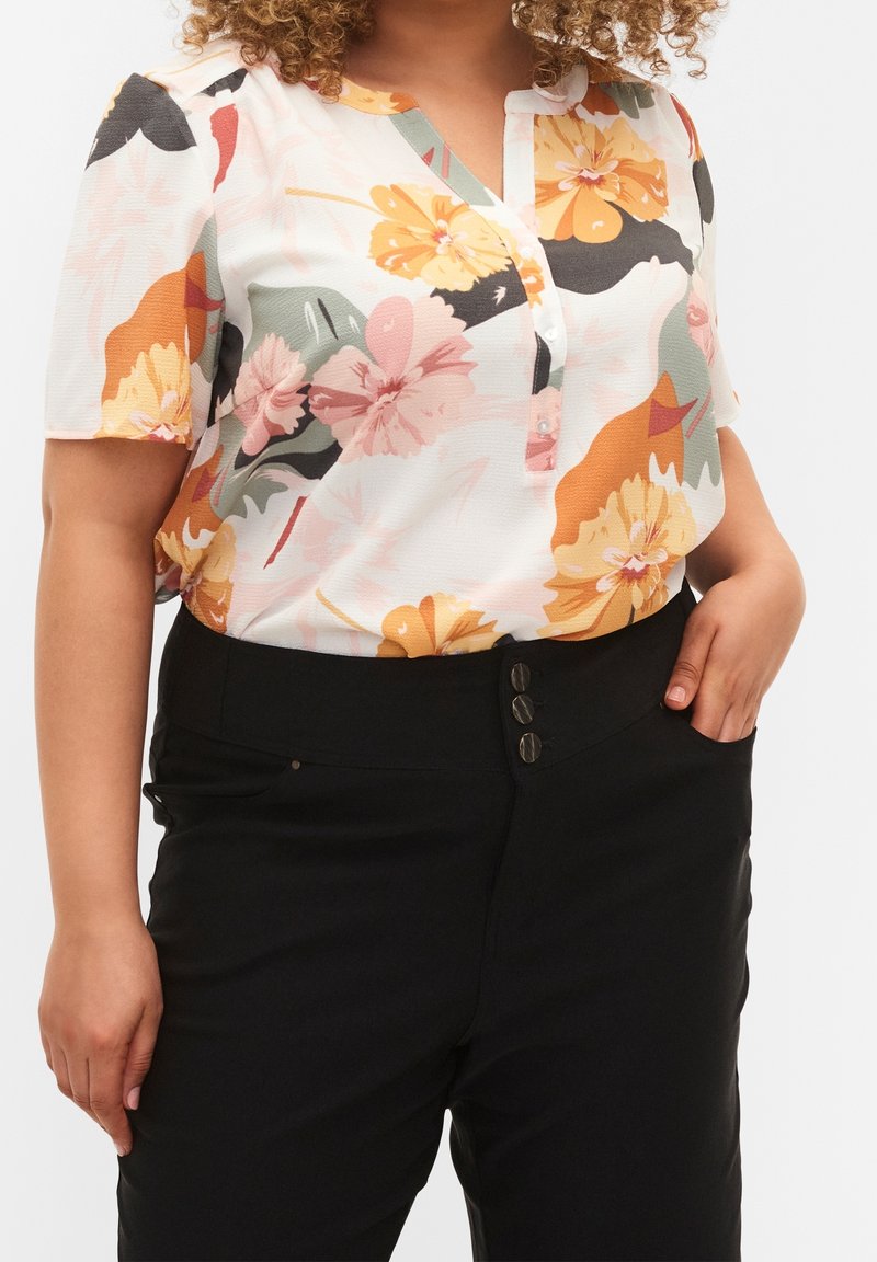 Femme portant un chemisier blanc à manches courtes à motifs floraux, rentré dans un pantalon noir taille haute avec trois boutons à l'avant.