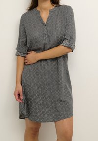Abito grigio con un motivo geometrico bianco, maniche arrotolabili e scollo henley. Il tessuto appare leggero e morbido.