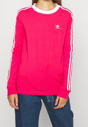 Person iført en lyserød Adidas langærmet skjorte med hvide striber på ærmerne og et lille Adidas-logo på brystet, kombineret med blå jeans.