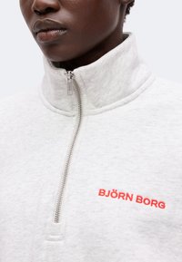 Gros plan d'une personne portant un haut zippé gris clair avec un col montant et un logo rouge "BJÖRN BORG" sur la poitrine.