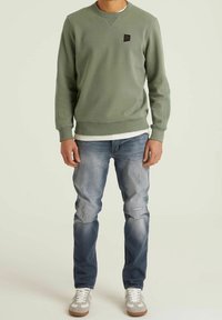 Sweat-shirt vert olive avec poignets et ourlet côtelés, arborant un petit patch logo noir. Associé à un jean bleu clair déchiré et des baskets décontractées.