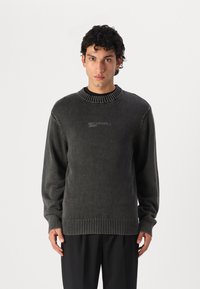 Mörkgrå stickad tröja med ribbad halsringning och mudd, med en subtil logotyp fram. Mjuk textur och avslappnad passform.