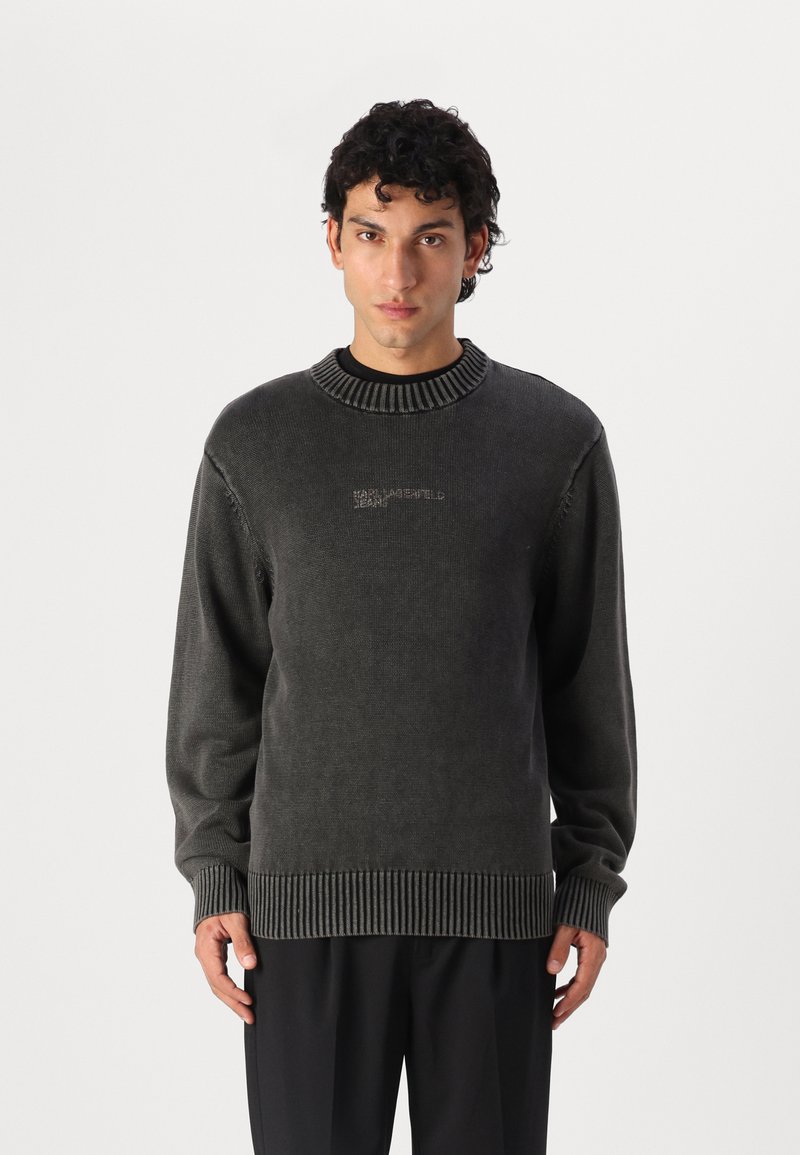 Mörkgrå stickad tröja med ribbad halsringning och mudd, med en subtil logotyp fram. Mjuk textur och avslappnad passform.