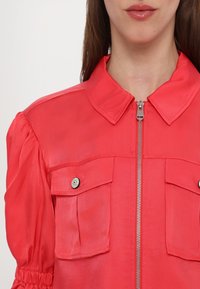 Punane lukuga jakk koos kaelusega, kahe rinnanäpiga ja kogutud varrukatega, mis lisavad tekstuuri. Sile kangas läikiva viimistlusega.