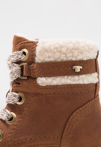 Bottine en suède marron avec un doublage crémeux et moelleux, œillets en métal et accent décoratif en métal sur le côté. Finition en cuir texturé.