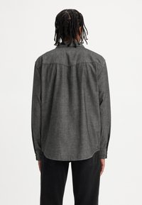 Camicia grigia a maniche lunghe con una texture liscia, collo abbottonato e orlo incurvato, con dettagli cuciti e vestibilità rilassata.