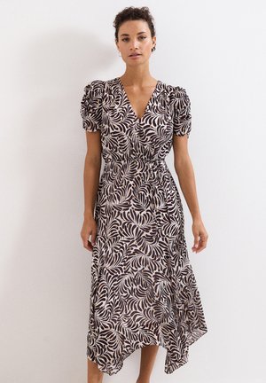 Woman wearing a V-neck, short puff sleeve dress with black and beige leaf patterns, standing against a white wall.