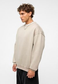 Beige oversized sweatshirt met verlaagde schouders, geribbelde manchetten en zoom; gemaakt van zacht, gestructureerd materiaal; gelaagd met een wit shirt eronder.