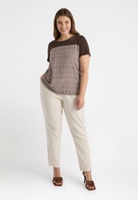 Top marrón de manga corta con patrón geométrico, pantalones crema a medida y sandalias de tacón con tiras marrones. La modelo está de pie con una mano en el bolsillo.