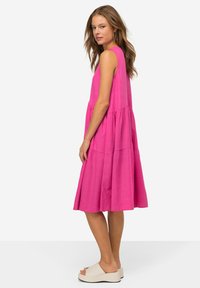 Abito di cotone rosa senza maniche con design a strati e lunghezza a metà polpaccio. Abbinato a sandali con la piattaforma di colore chiaro. Sfondo semplice.