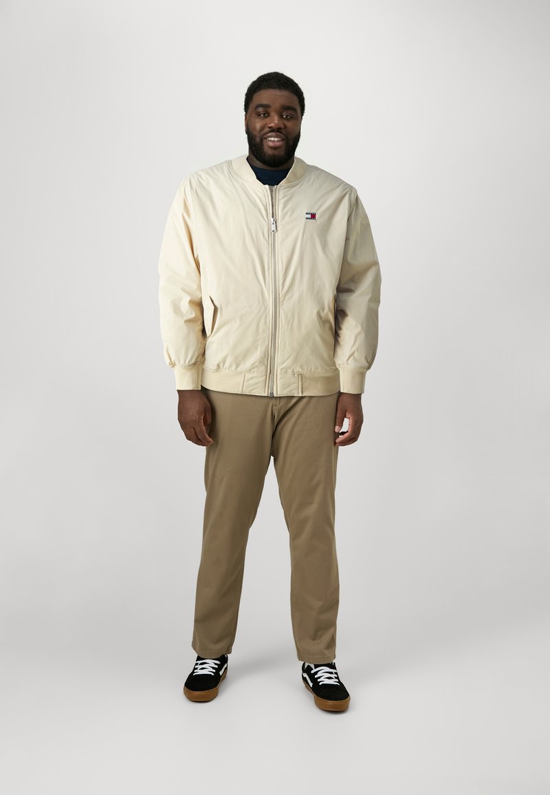Lysebeige bomberjakke med glidelås foran, ribbede mansjetter og Tommy Hilfiger-logo. Kombinert med khakibukser og svarte joggesko.